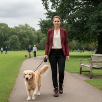 A woman walks a golden retriever on a paved path in a park, wearing a burgundy blazer, white top, black pants, and dark brown boots.