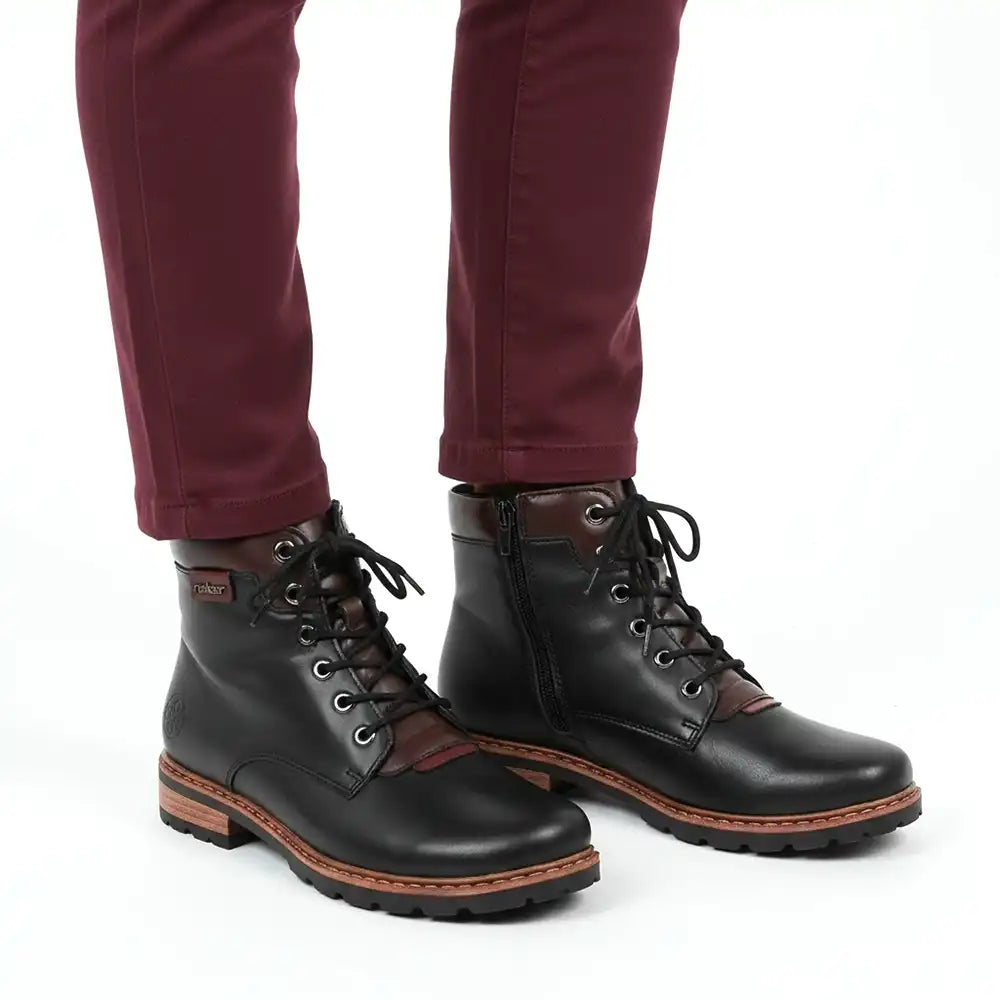 A pair of women's black leather lace-up boots with brown side panels and brown stitching, shown on a model wearing burgundy pants against a white background.