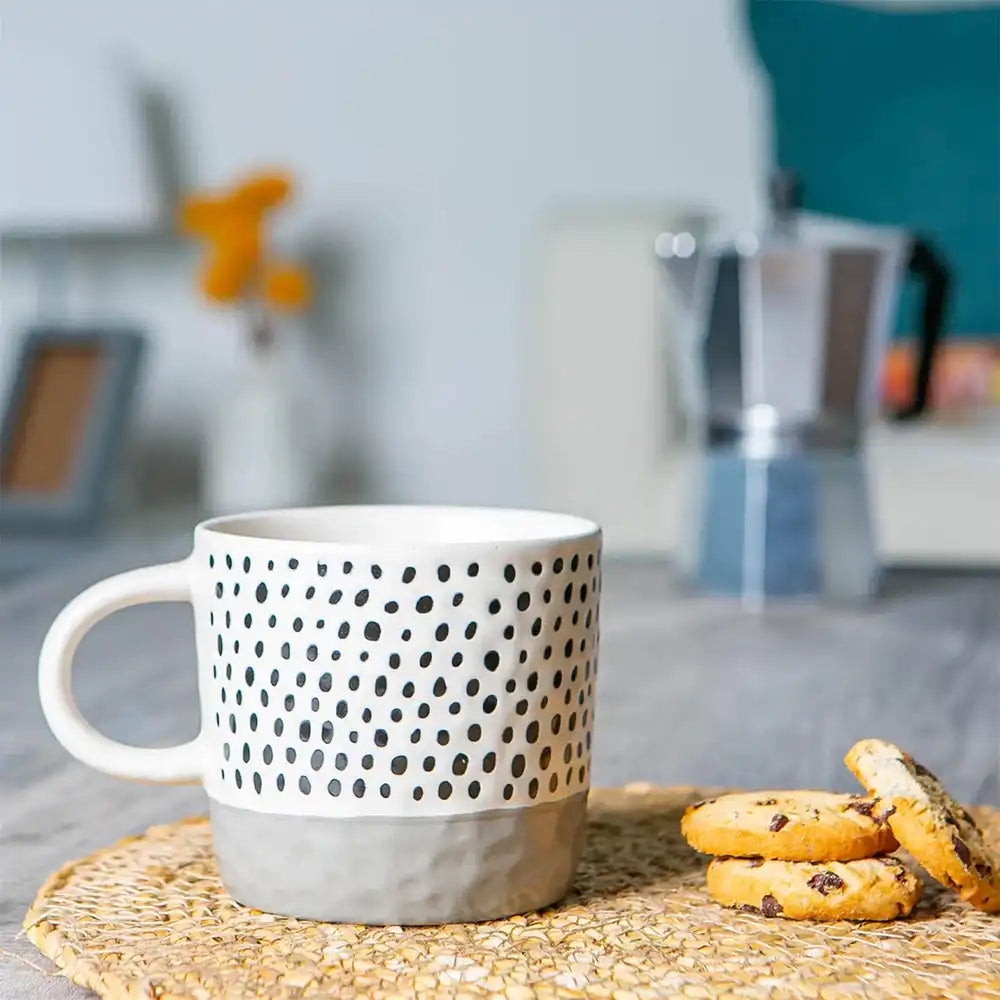 White mug with black dot pattern on a woven coaster with cookies, blurred kitchen background