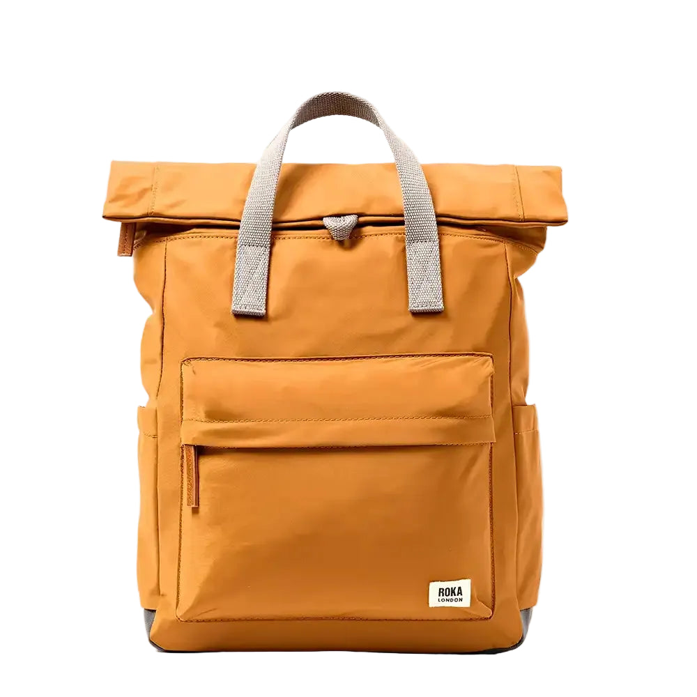 A caramel roll-top backpack with gray webbed handles and a small white 'ROKA' brand patch on the lower front, shown against a plain white background.