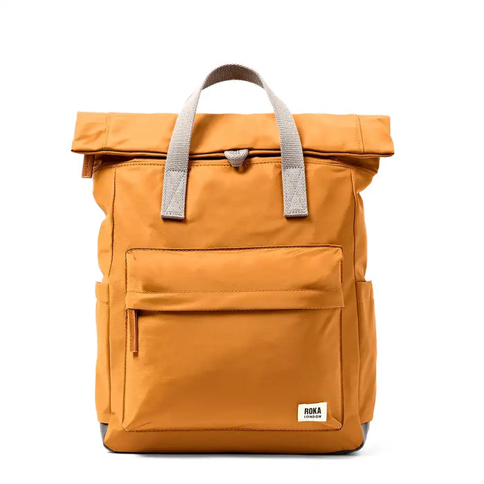 A caramel roll-top backpack with gray webbed handles and a small white 'ROKA' brand patch on the lower front, shown against a plain white background.