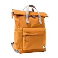 Side view of a caramel backpack with roll top and front pocket with ROKA label in white on bottom right corner and a slip pocket on the side.