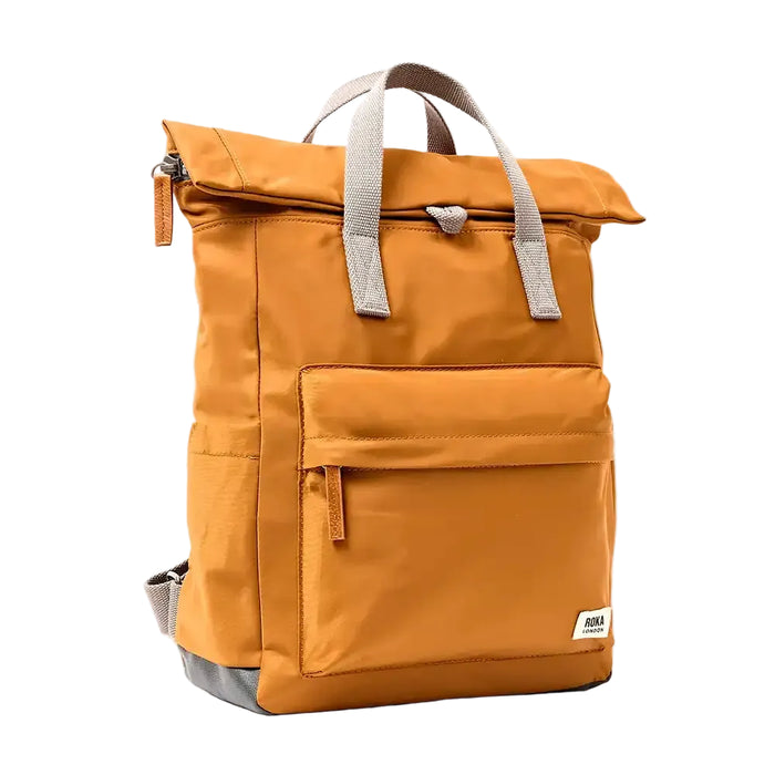 Side view of a caramel backpack with roll top and front pocket with ROKA label in white on bottom right corner and a slip pocket on the side.