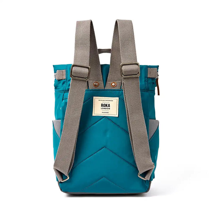 A back view of a teal blue and grey ROKA backpack shown from the back, featuring padded grey shoulder straps, a cream brand label, and a quilted chevron pattern on a white background.
