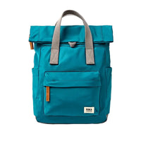 Front view of a teal blue roll-top backpack with grey webbing handles and a front pocket, featuring a small white 'ROKA' brand label on the lower right corner.