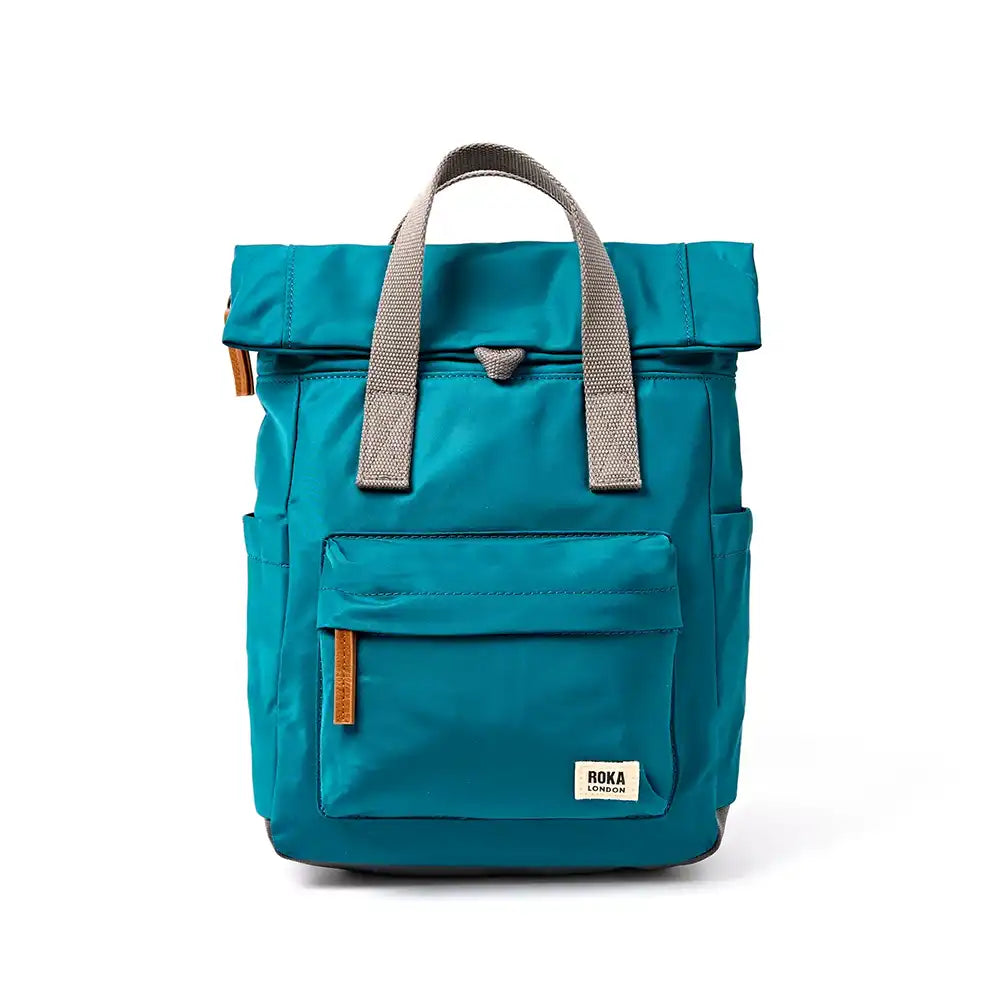 A teal blue roll-top backpack with grey webbing handles and a front pocket, featuring a small white 'ROKA' brand label on the lower right front, photographed against a clean white background.
