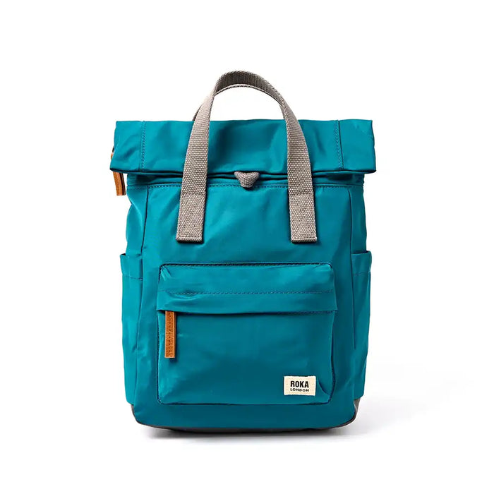 A teal blue roll-top backpack with grey webbing handles and a front pocket, featuring a small white 'ROKA' brand label on the lower right front, photographed against a clean white background.