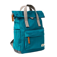 Side view of a teal blue roll-top backpack with grey webbed handles, grey bottom trim, and small brown leather accents on the zipper pulls and a front patch, shown on a plain white background.