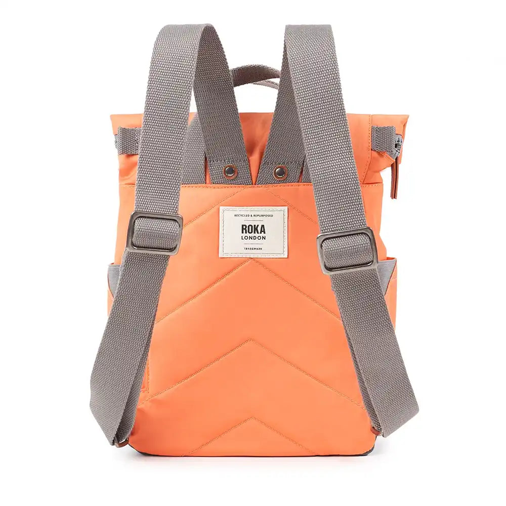 A back view of an orange roll-top backpack with grey webbing shoulder straps and a white rectangular brand patch that reads 'ROKA LONDON'.