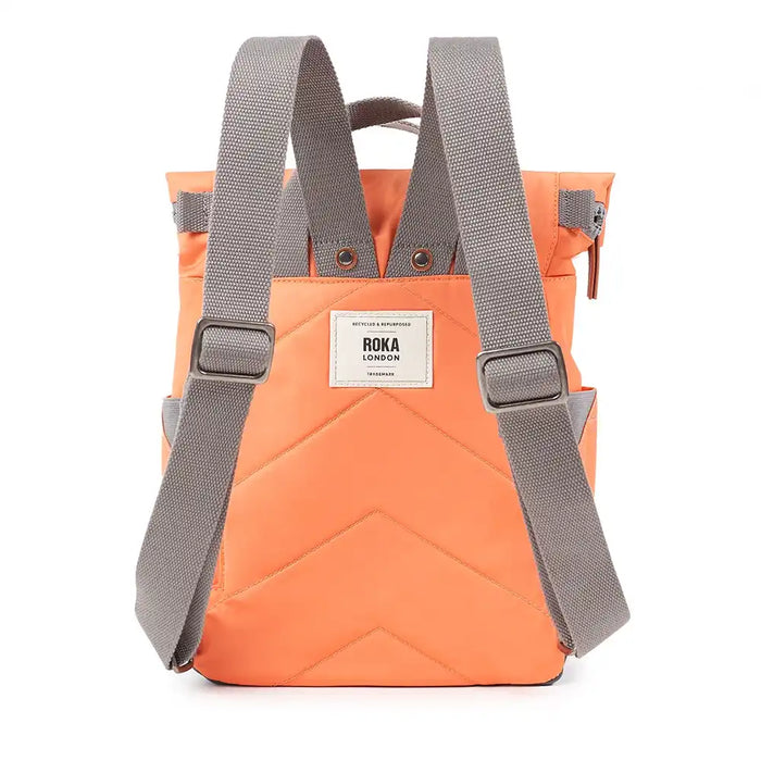 A back view of an orange roll-top backpack with grey webbing shoulder straps and a white rectangular brand patch that reads 'ROKA LONDON'.