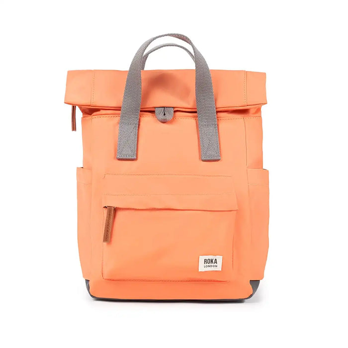 An orange roll-top backpack with grey webbed handles and a small white ROKA London brand patch on the front pocket, shown against a plain white background.