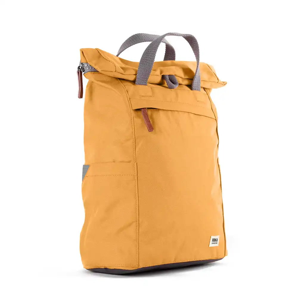 Side view of a yellow backpack with roll top and front pocket with ROKA label in white on bottom right corner and a slip pocket on the side.