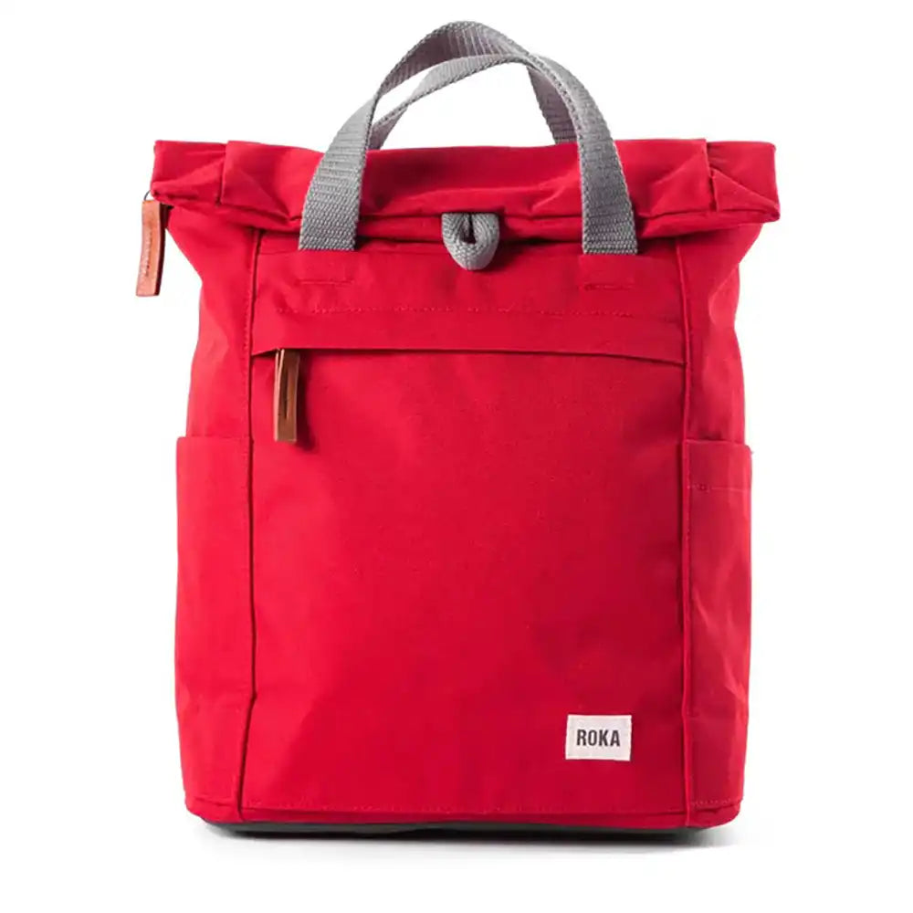 A red roll-top backpack with grey webbed handles, side pockets, and a small white ROKA brand label on the lower front, photographed on a clean white background.