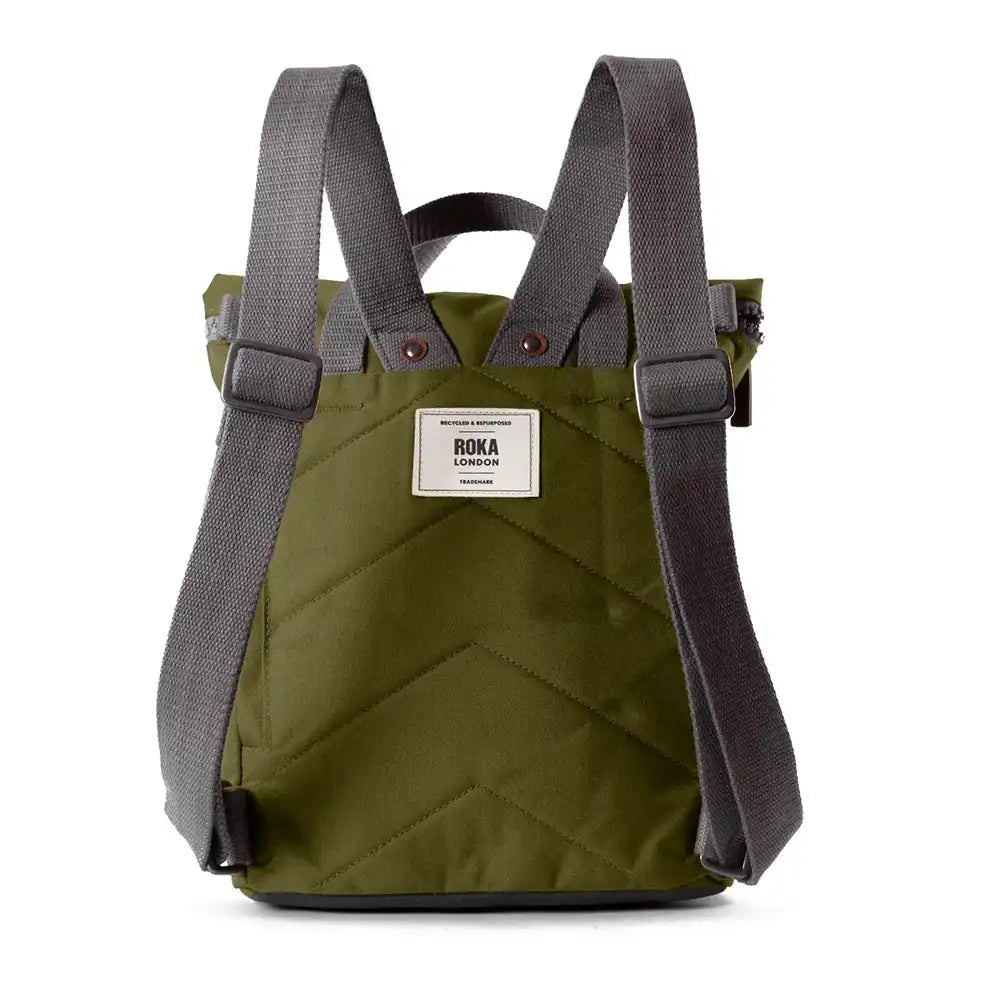A back view of a dark olive green quilted canvas backpack with dark grey adjustable shoulder straps and a white brand label on the front, shown against a plain white background.