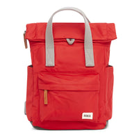Front view of a cranberry red backpack with roll top and front pocket with ROKA label in white on bottom right corner.