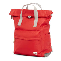 Side view of a cranberry red backpack with roll top and front pocket with ROKA label in white on bottom right corner and a slip pocket on the side.