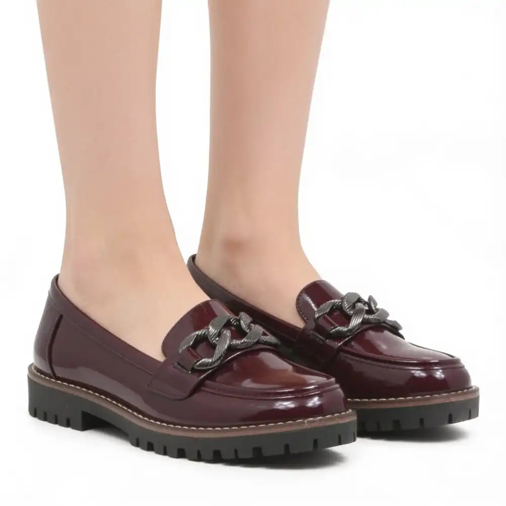 A pair of glossy burgundy patent leather penny loafers with a dark metal chain detail across the vamp and chunky black lug soles, shown on a model's feet against a white background.