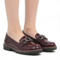 A pair of glossy burgundy patent leather penny loafers with a dark metal chain detail across the vamp and chunky black lug soles, shown on a model's feet against a white background.