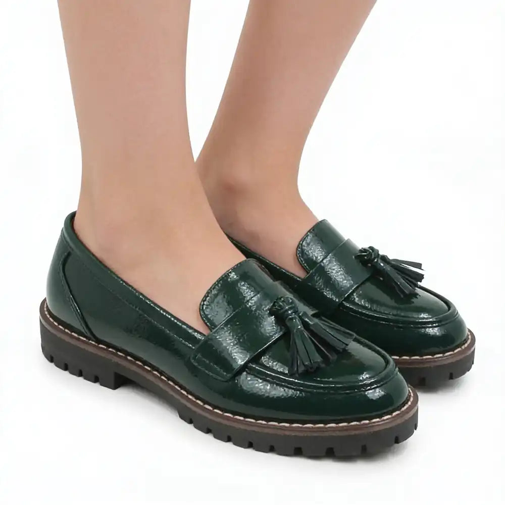 A pair of dark green patent leather tassel loafers with chunky brown and black lug soles, shown on a model's feet against a white background.