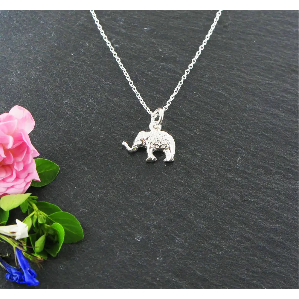 Silver elephant pendant necklace on a dark surface with pink flowers and green leaves.