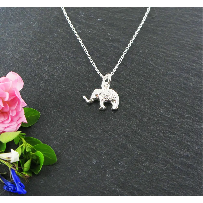 Silver elephant pendant necklace on a dark surface with pink flowers and green leaves.