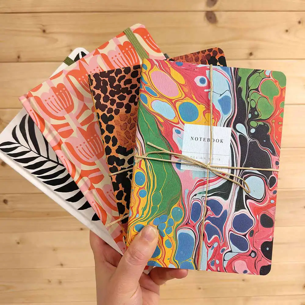 A hand holds a fan of four colorful patterned notebooks with elastic-wrapped covers, set against a light wood plank background. The notebooks feature marbled, floral, and leopard-style designs in bright colors, with a small white label reading 'NOTEPAD' on the front.
