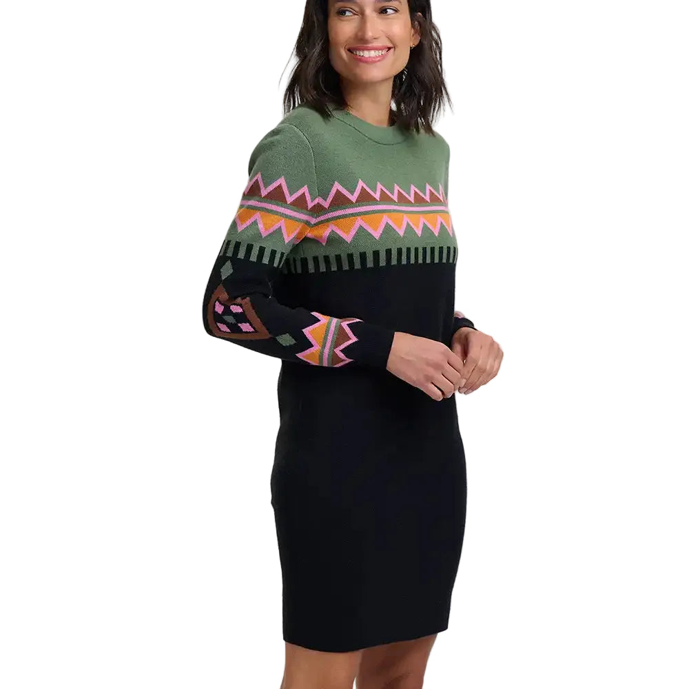 A woman wearing a long-sleeve knit sweater dress with a black body, olive green yoke, and a colorful geometric pattern across the chest and sleeves, photographed against a light gray studio background.