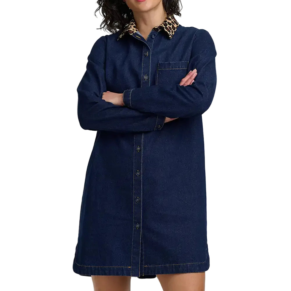A model wearing a dark blue denim shirt dress with a contrasting leopard-print collar, standing with arms crossed against a light gray background.