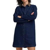 A model wearing a dark blue denim shirt dress with a contrasting leopard-print collar, standing with arms crossed against a light gray background.