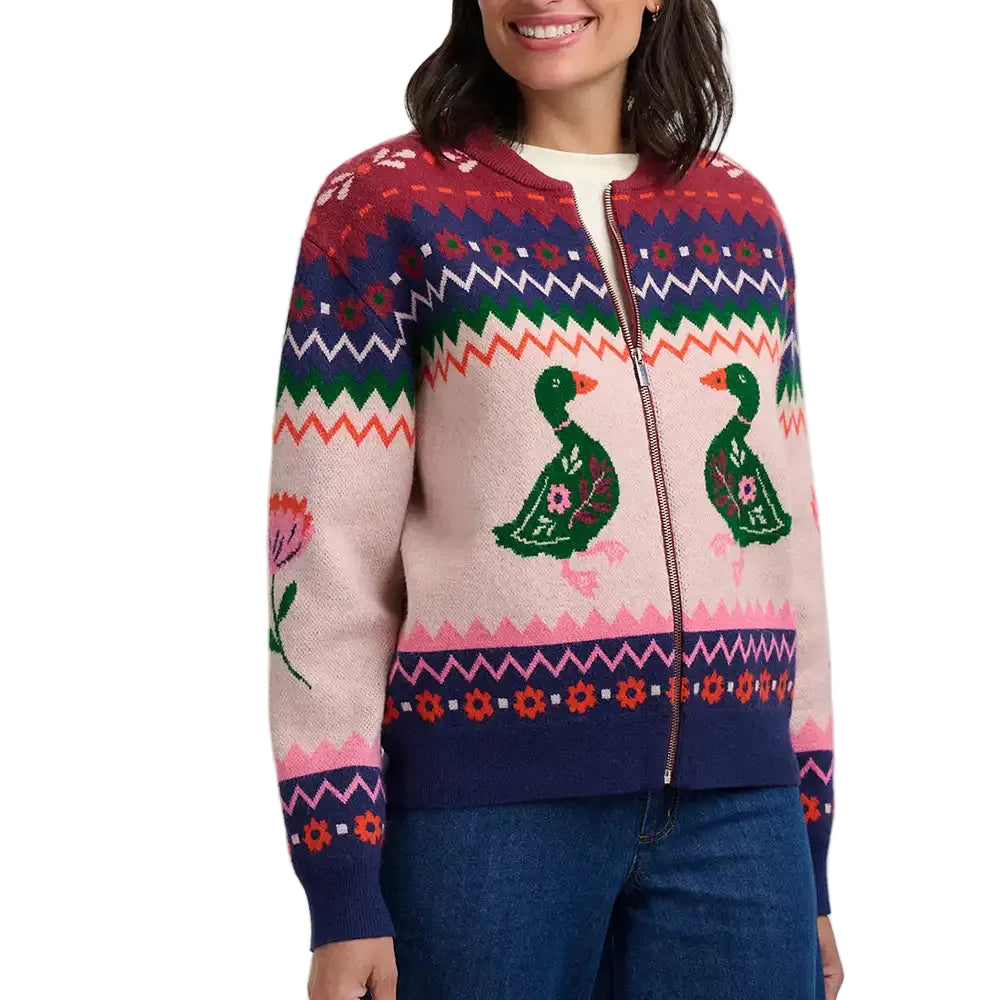 A woman wearing a colorful knit zip-up sweater with Fair Isle patterns, featuring green ducks and floral motifs on a light pink base, paired with blue jeans against a plain light gray background.