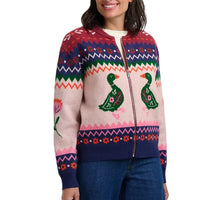 A woman wearing a colorful knit zip-up sweater with Fair Isle patterns, featuring green ducks and floral motifs on a light pink base, paired with blue jeans against a plain light gray background.