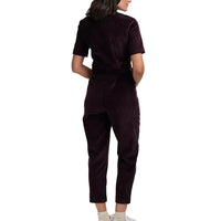 Woman wearing a short sleeved, cropped leg dark plum coloured, zip-up  boilersuit in a white studio environment. She has her back to the camera.