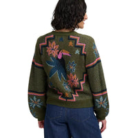 Woman wearing khaki green four button cardigan with flowers and toucans pattern. She is in a white studio environment. She has her back to the camera.