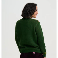 A woman shown from the back wearing a dark green knit sweater with subtle gold and pink patterned detailing on the shoulders and cuffs, paired with dark corduroy pants against a light gray studio background.