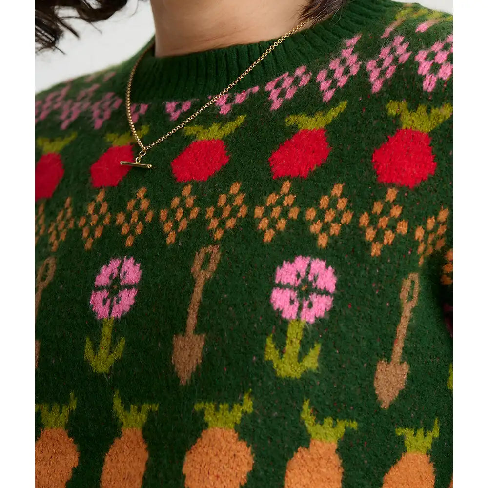 A close-up of a person wearing a dark green knit sweater with a colorful pattern of red strawberries, pink flowers, orange pumpkins, and brown gardening tools, accessorized with a gold necklace.