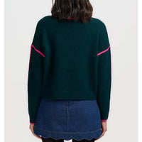 A person shown from the back wearing a dark green knit sweater with bright pink contrast trim along the shoulders and cuffs, paired with a blue denim skirt, against a light gray studio background.