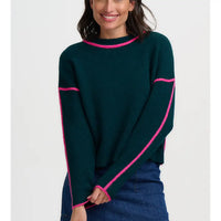 A woman wearing a dark green knit sweater with bright pink contrast trim along the crew neck, cuffs, hem, and across the upper arms, paired with a blue denim skirt against a plain light gray background.
