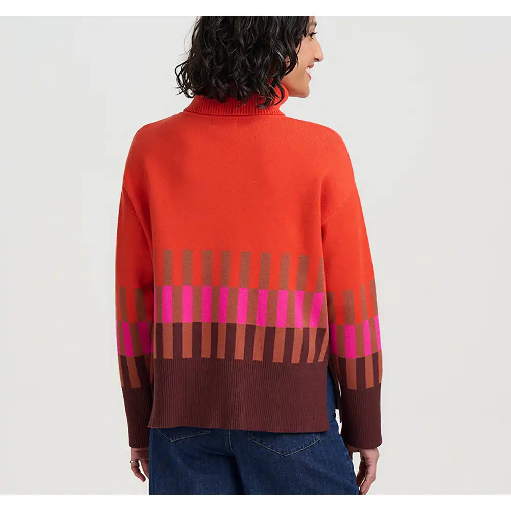 A woman shown from the back wearing a bright orange-red knit sweater with a brown ribbed turtleneck and a geometric stripe pattern in brown, pink, and orange across the lower back and sleeves, paired with blue jeans against a light gray studio background.