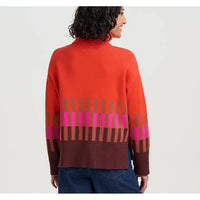 A woman shown from the back wearing a bright orange-red knit sweater with a brown ribbed turtleneck and a geometric stripe pattern in brown, pink, and orange across the lower back and sleeves, paired with blue jeans against a light gray studio background.