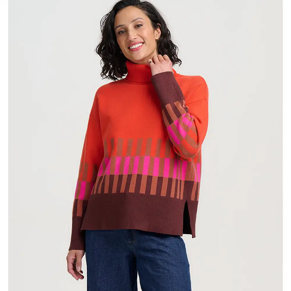 A woman modeling a bright orange turtleneck sweater with a brown and pink geometric stripe pattern across the chest and sleeves, paired with blue jeans against a white background.