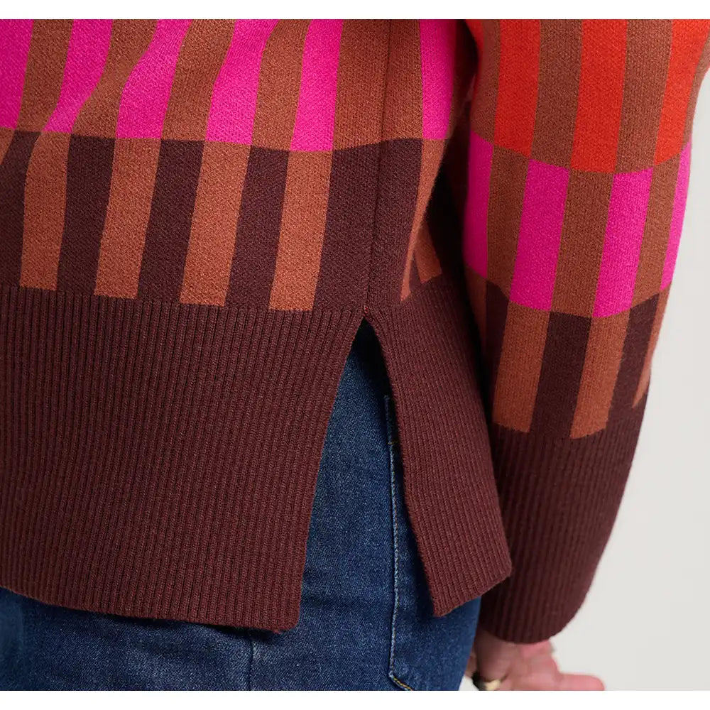 Close-up of a ribbed knit sweater with a geometric plaid pattern in brown, terracotta, and bright pink, worn over blue jeans.
