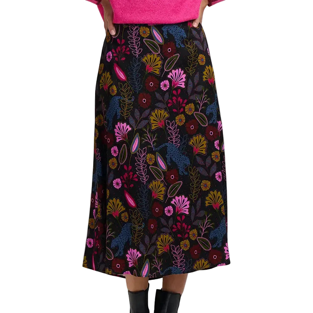 A black midi skirt with a vibrant, multicolored botanical pattern featuring pink, yellow, blue, and red floral and leaf motifs, shown on a model wearing a hot pink top and black boots.