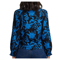 A back view of a woman wearing a long-sleeve blouse with a vibrant blue floral and bird pattern on a black background.