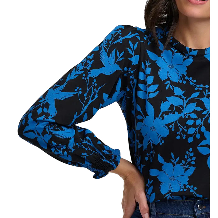 View of the arm of a woman wearing a round neck top with a blue pattern of hummingbirds and flowers on a black background.
