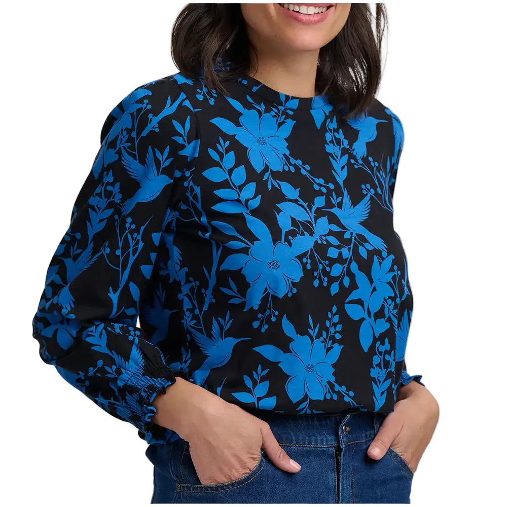 A woman wearing a long-sleeve blouse with a vibrant blue floral and bird pattern on a black background, paired with blue jeans against a plain white studio background.