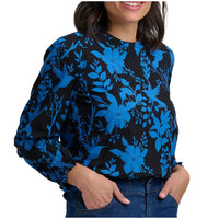 A woman wearing a long-sleeve blouse with a vibrant blue floral and bird pattern on a black background, paired with blue jeans against a plain white studio background.
