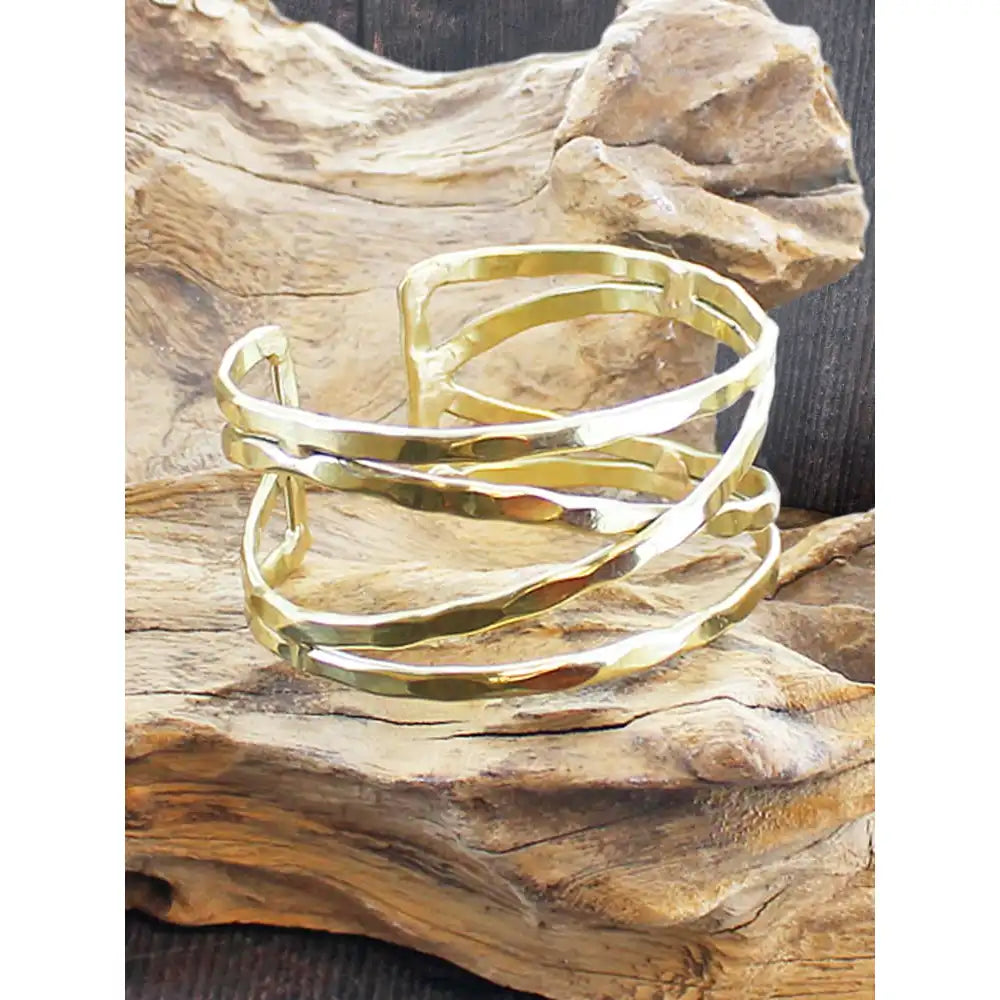 A polished gold-toned metal cuff bracelet with a hammered texture, displayed on a piece of natural driftwood.