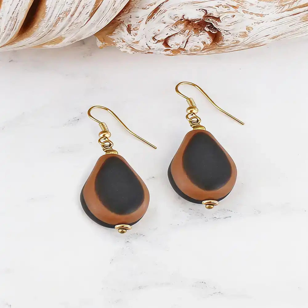 A pair of teardrop-shaped drop earrings with black beads bordered in brown, hanging from gold fishhook wires on a white marble surface with seashells in the background.
