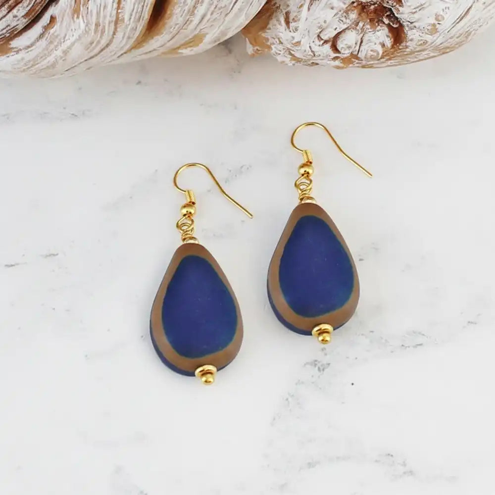 A pair of teardrop-shaped drop earrings with deep blue beads bordered in brown, hanging from gold fishhook wires on a white marble surface with seashells in the background.