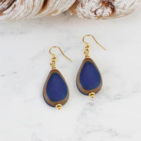 A pair of teardrop-shaped drop earrings with deep blue beads bordered in brown, hanging from gold fishhook wires on a white marble surface with seashells in the background.